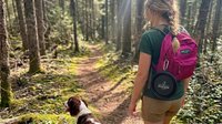 Person hiking with a dog in a forest, wearing a pink backpack with a visible brand logo.